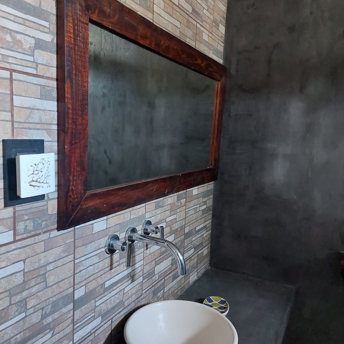 A modern bathroom is featured with textured stone walls and a large wooden-framed mirror above a circular basin sink. A stylish faucet and minimalist design elements enhance the space, creating a clean and functional atmosphere.