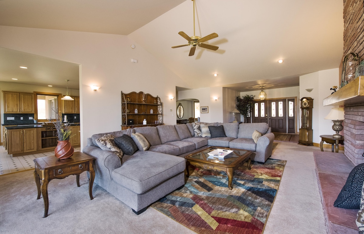 A spacious living area features a large sectional sofa, complemented by a decorative coffee table atop an area rug with various colors. Soft lighting enhances the room's warmth, and a ceiling fan provides air circulation. The open layout connects to the kitchen and entryway.