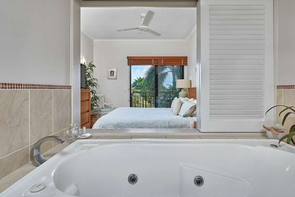 A spa bath is positioned in the foreground, offering a view through a window into a serene bedroom featuring a spacious bed and natural light. Lush greenery is visible outside, with soft furnishings enhancing the tranquil ambiance.