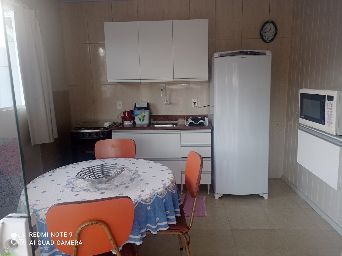 A compact kitchen area is presented, featuring a round dining table with orange chairs and a blue tablecloth. A white refrigerator and microwave are positioned alongside a stove and countertop. Natural light enters through a window, complementing the neutral color scheme of the space.