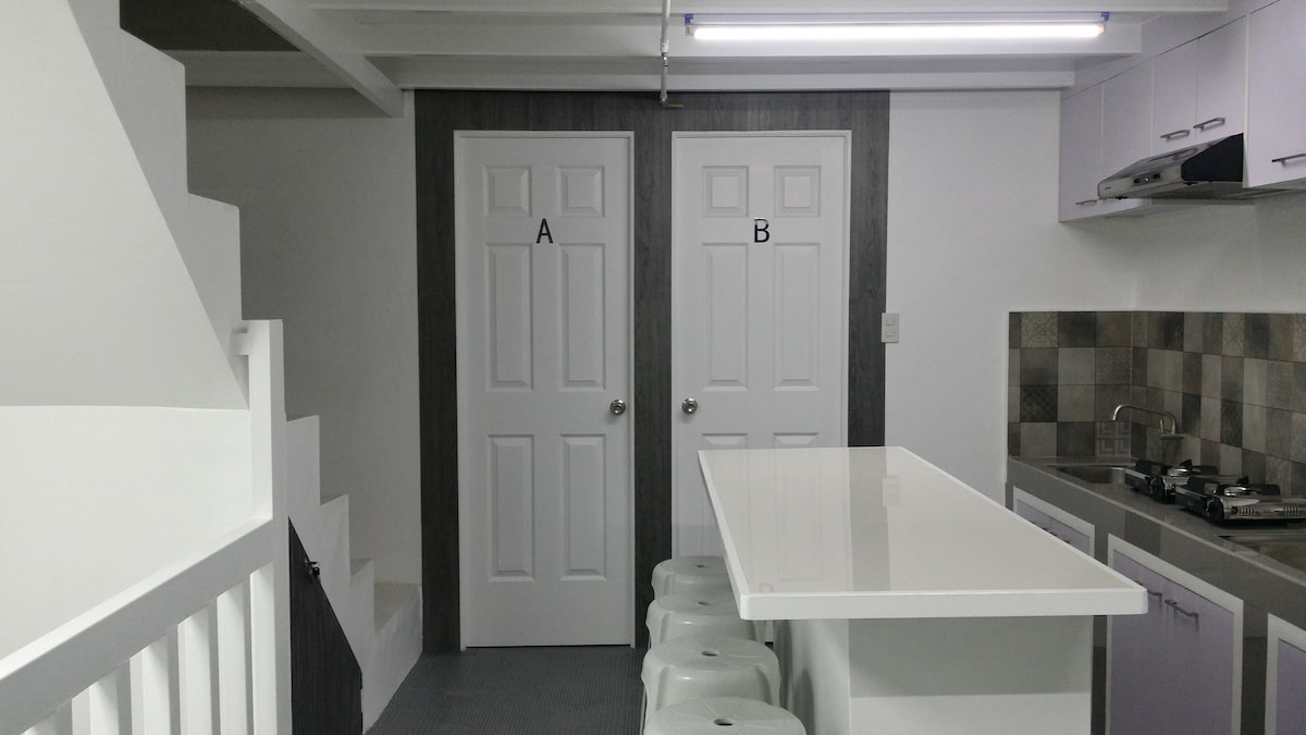 A modern kitchen area features a sleek white countertop with three stools, surrounded by cabinetry in muted tones. Two labeled doors, A and B, stand side by side against a gray wall. A staircase ascends to a second level, adding depth to the open layout.