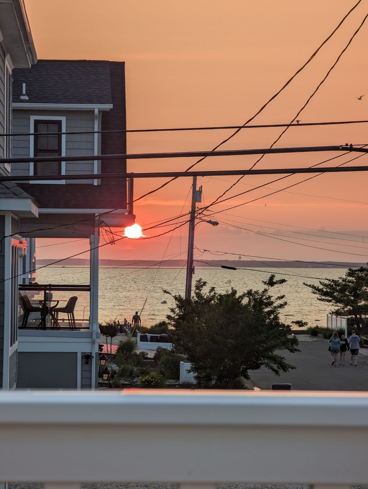 A sunset casts warm colors across the sky, visible over the water. The scene includes a view of nearby homes and silhouettes of people walking along the shoreline. Power lines and trees frame the image, adding depth to the coastal atmosphere.