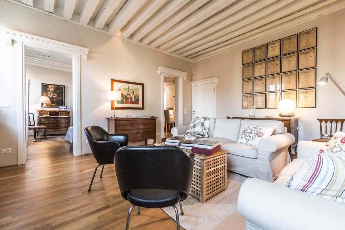 The living room features light wooden beams across a high ceiling and a mix of comfortable seating, including a pair of black chairs and a light-colored sofa. A wooden side table and decorative wall art contribute to a welcoming ambiance. Natural light fills the space.