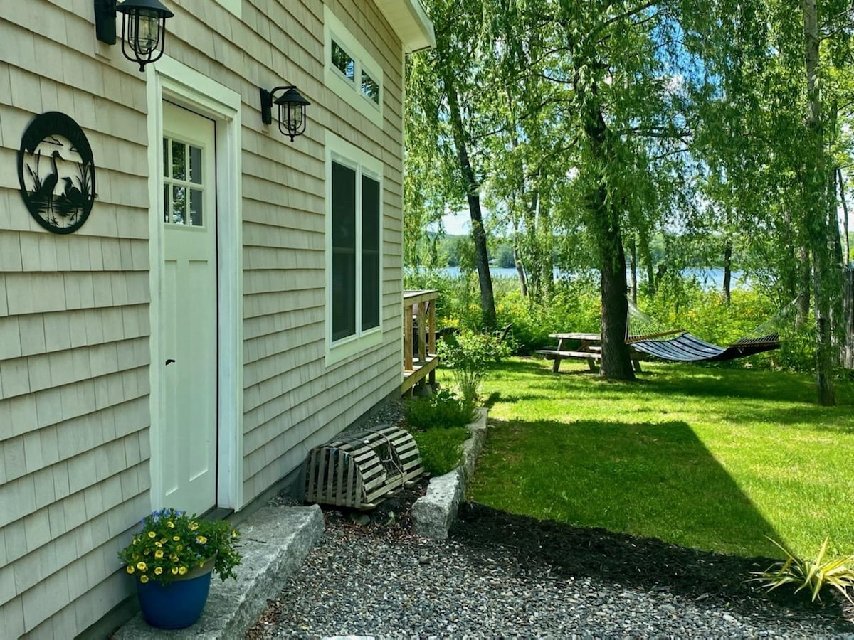 The exterior showcases the cottage's entrance, framed by light brown shiplap siding. A blue flower pot with vibrant blooms is positioned near the door. A grassy area extends to the lake, featuring a hammock and wooden picnic table under the trees, creating a serene outdoor space.