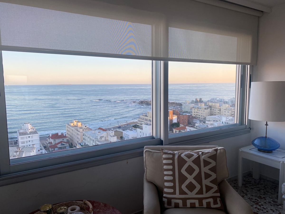 A large window offers a panoramic view of the sea and coastline, with gentle waves meeting the shore. The interior features a cozy chair, a patterned throw pillow, and a lamp, enhancing the tranquil setting.