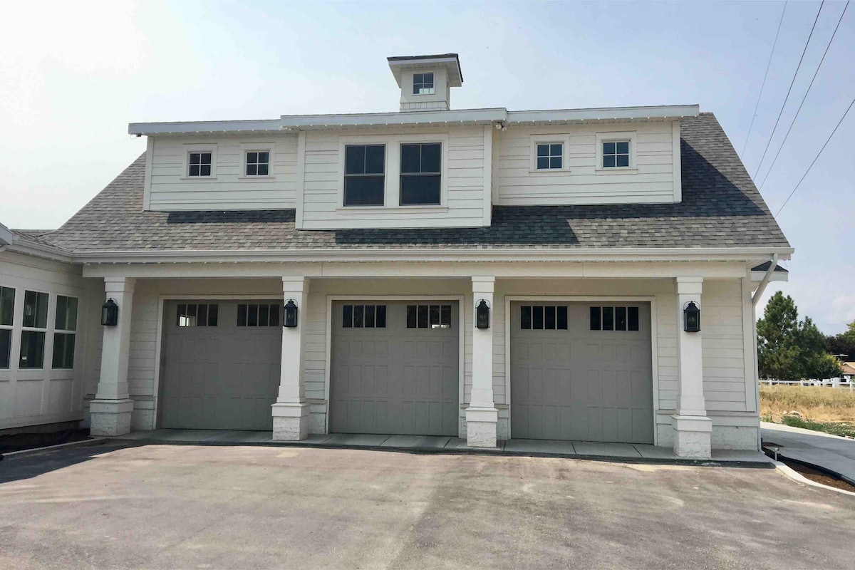 The exterior of the carriage house presents a classic design, featuring a three-car garage with large doors and multiple windows. The well-maintained façade is accented by a small cupola at the roofline, framed by a clear blue sky.