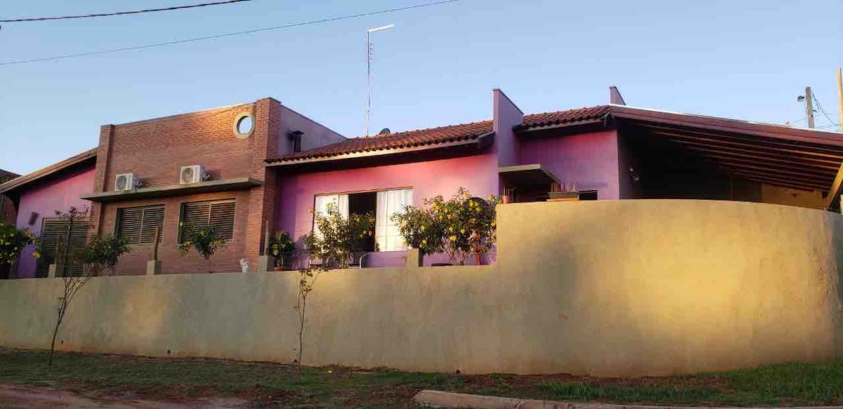 A one-story house is presented with a blend of purple and brick exteriors. Air conditioning units are visible on the front wall. Tall windows allow natural light inside, framed by flowering plants, while a low wall adds privacy to the property.