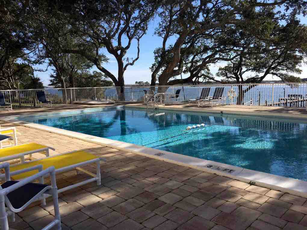 A clear blue pool is surrounded by a spacious stone patio featuring yellow lounge chairs. Mature trees provide natural shade, and the calm water of the scenic sound is visible in the background. A safety fence encloses the pool area.