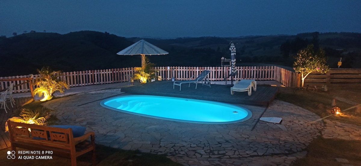 A softly lit swimming pool glows in turquoise hues under a twilight sky, surrounded by landscaped gardens. Lounge chairs are positioned nearby, along with a shaded gazebo. The distant hills are visible, creating a serene backdrop for relaxation.