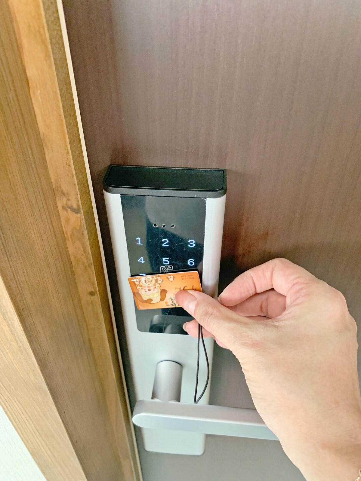 A keyless entry system is shown on a wooden door, featuring a numeric keypad. A hand is seen holding a card, poised to swipe it near the reader. The door's material has a natural wood finish.