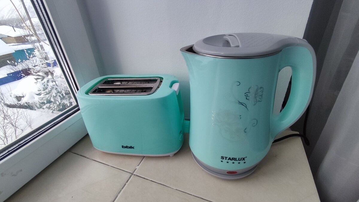 A turquoise kettle and toaster are positioned on a countertop by a window. The kettle features a subtle floral design, and both appliances are designed to complement each other in color. Outside, snow can be seen through the window, enhancing a cozy environment.