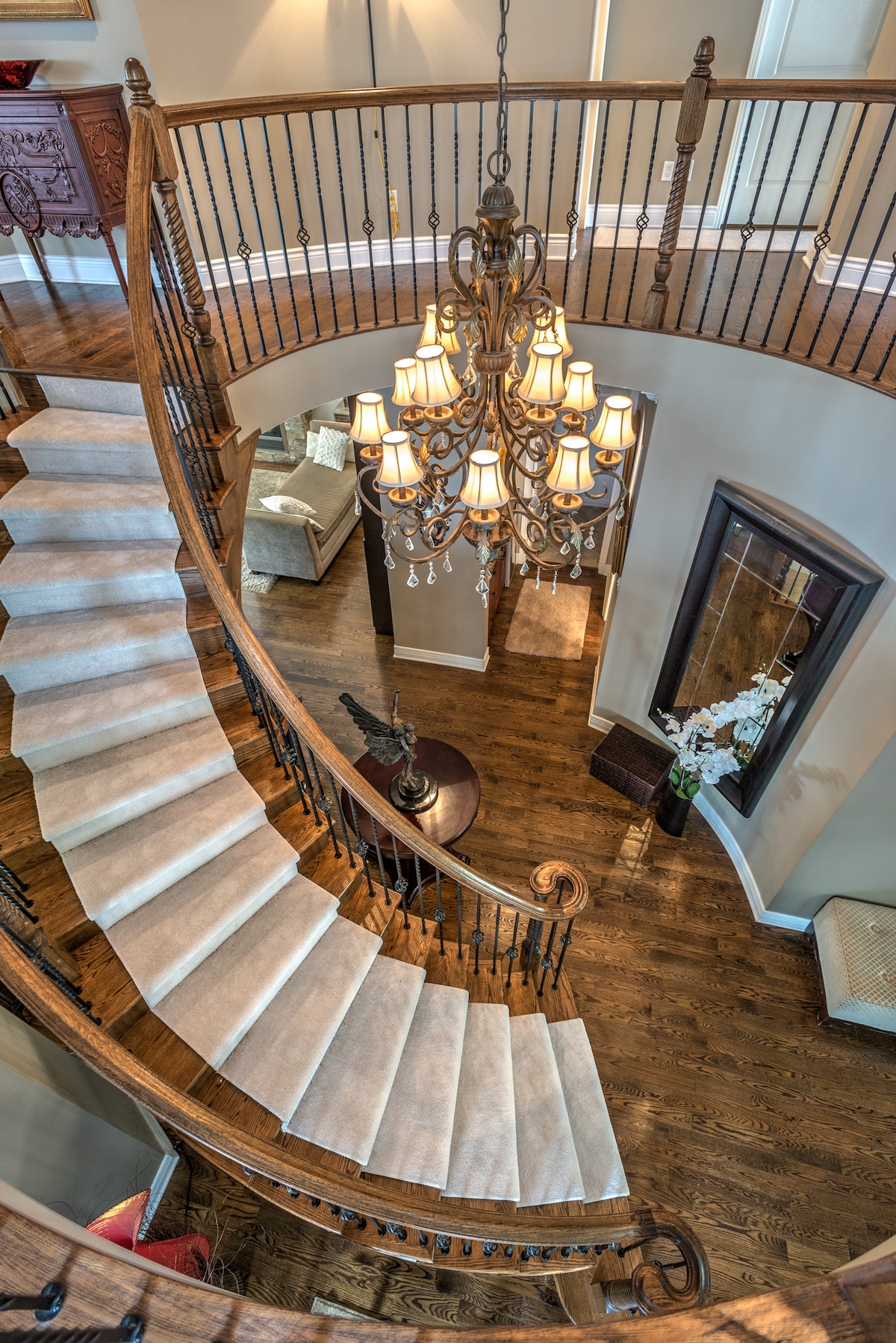 A grand staircase is featured, winding elegantly with soft beige carpeting. A chandelier hangs from the ceiling, illuminating the space below. A mirror and decorative table accentuate the landing area, while plush seating is visible in a nearby room, creating a warm and welcoming ambiance.