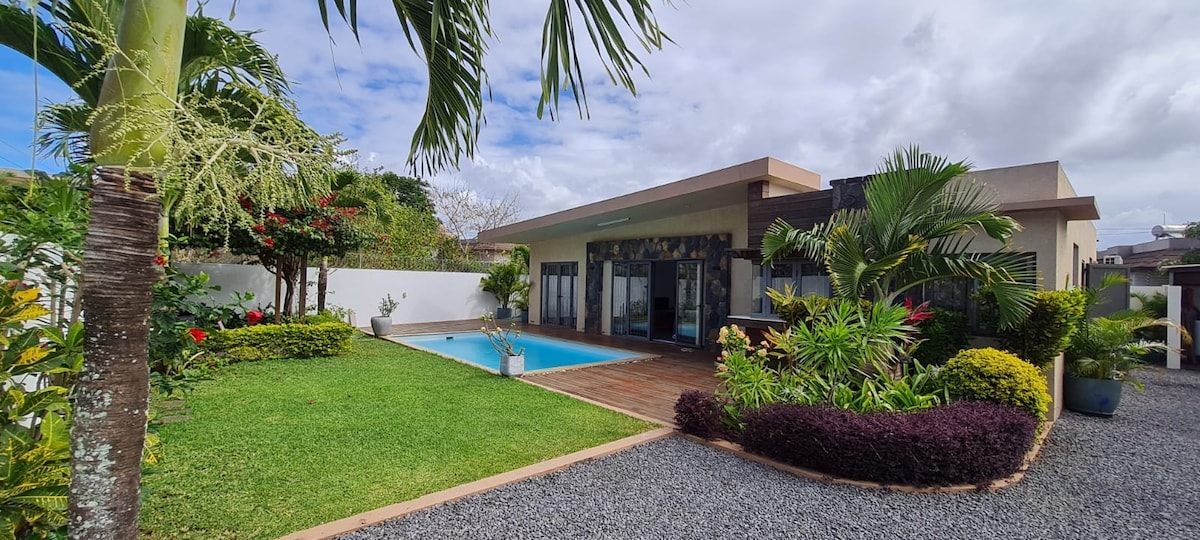 The modern villa is surrounded by tropical landscaping, with a private pool visible in the foreground. Lush greenery frames the space, while a wooden deck complements the outdoor area. Large glass doors open to the living space, creating a harmonious blend of indoor and outdoor environments.