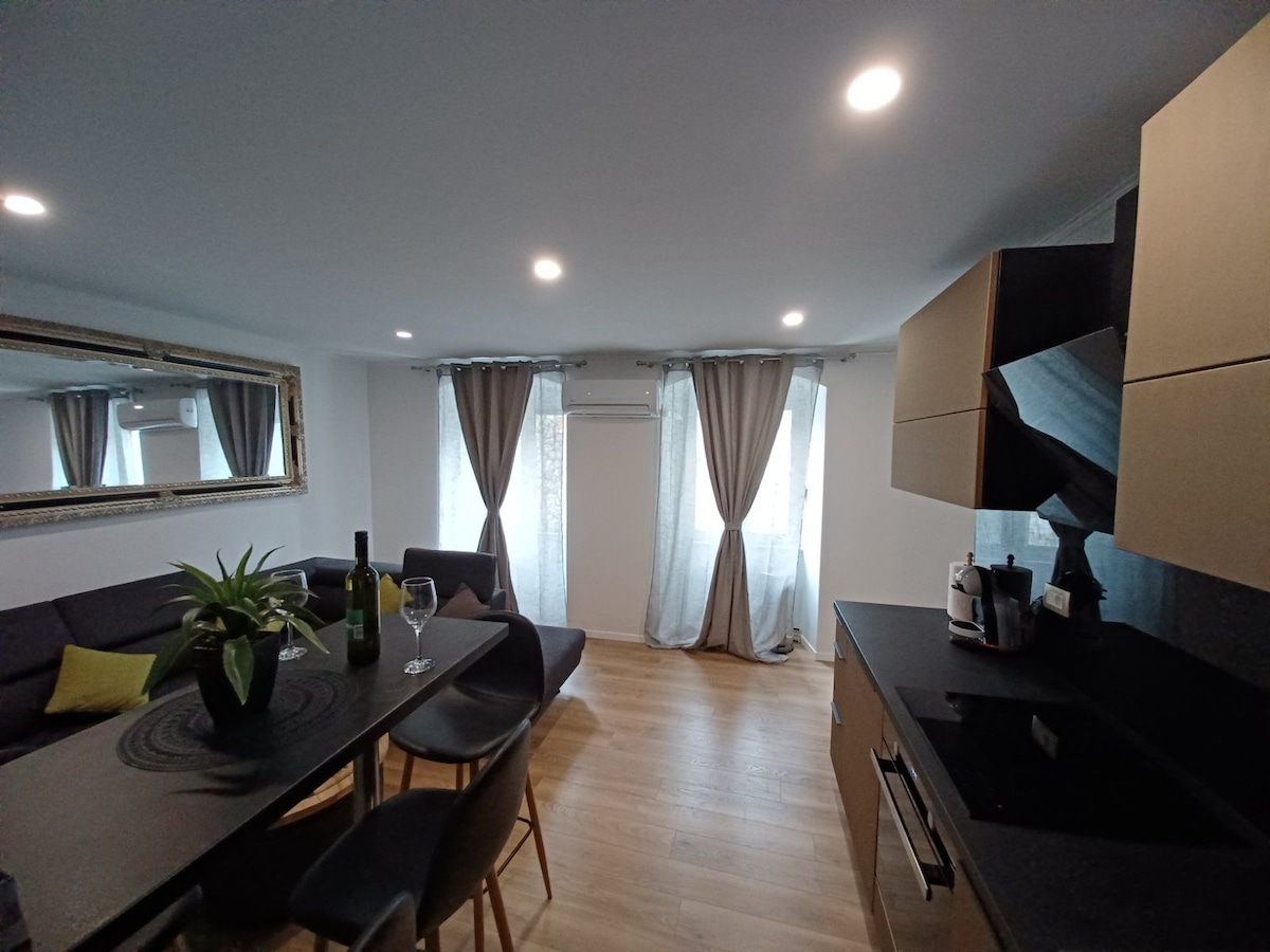 A modern living area features a dark sofa and a wooden dining table, both enhancing the space's functionality. Natural light enters through large windows, adorned with light gray curtains. A mirror reflects the room, while plants add a touch of greenery to the decor.