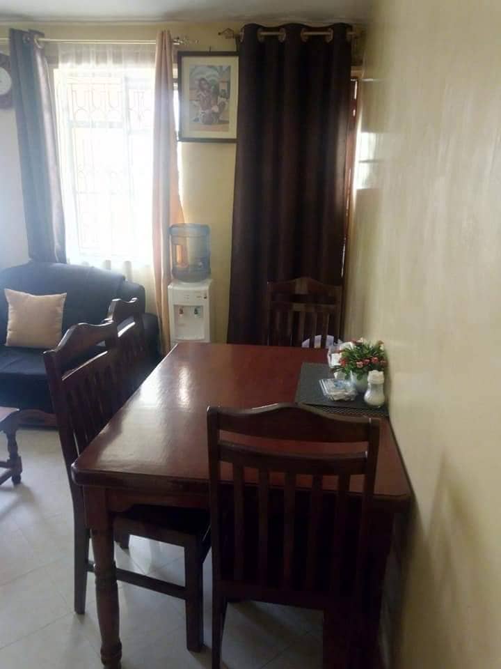 A cozy dining area features a wooden table surrounded by four chairs. A small vase with flowers rests on a table mat, alongside condiments. Natural light enters through a window, highlighting a nearby water dispenser and a dark-colored couch against the wall.