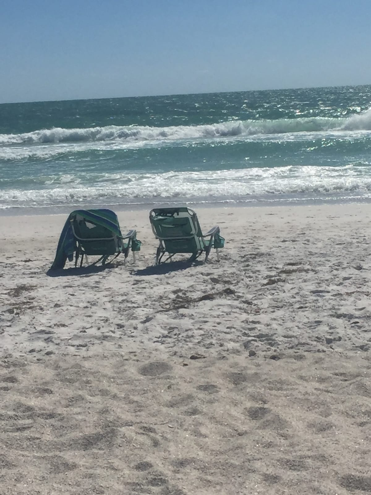 Two beach chairs are positioned on a sandy shore, facing the ocean. Gentle waves can be seen lapping at the beach, while the sun casts a bright light over the water, creating a serene coastal environment.