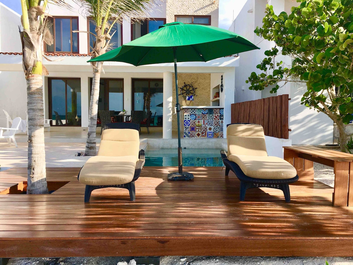 A wooden terrace features two lounge chairs under a green umbrella, overlooking a serene swimming pool. In the background, large glass doors provide a view into the indoor living space, while tropical plants enhance the inviting ambiance.
