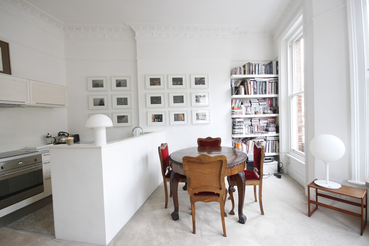 The spacious dining area features a round table surrounded by four chairs, combining modern and classic design elements. A bookshelf filled with books stands against the wall, while large windows allow natural light to brighten the room. The kitchen area is partially visible with contemporary appliances.