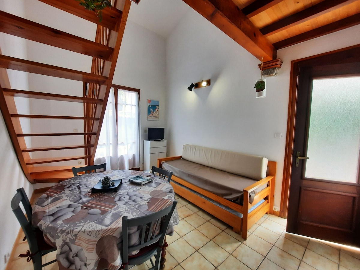 An open-plan living area is featured, showcasing a light-colored futon and a round dining table set with decorative items. Natural light floods the space through a window adjacent to the door leading outside. A staircase leads to the upper level, enhancing the room's spacious feel.