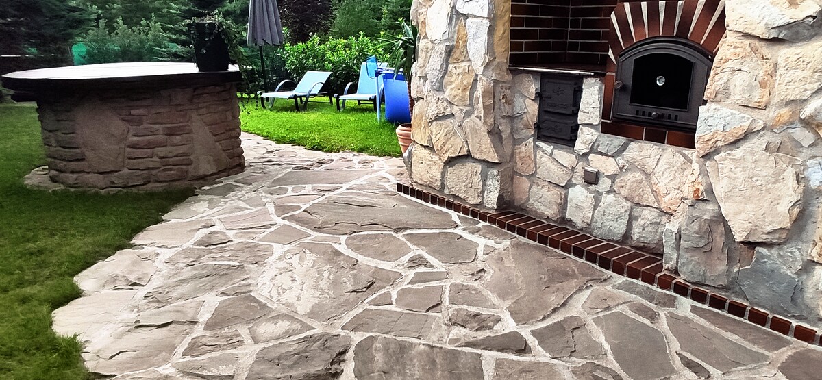 A stone pathway leads through a landscaped yard, connecting a seating area and an outdoor grill. A round stone table is positioned nearby, with lounge chairs placed in the background, surrounded by greenery.