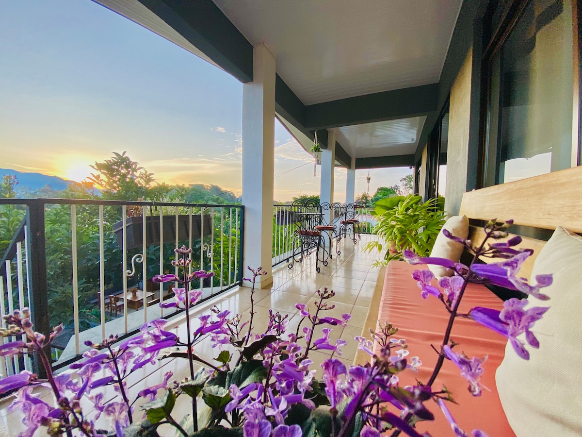 A spacious terrace is showcased, adorned with vibrant purple flowers and potted plants. Comfortable seating arrangements are positioned against the railing, providing views of the surrounding greenery and sunset. The tranquil environment is highlighted by the soft light of dusk.