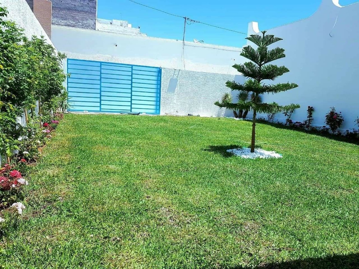 A well-maintained garden features lush green grass and a small tree at its center, surrounded by flower beds. A blue gate is visible in the background, contrasting with the white wall that encloses the area.