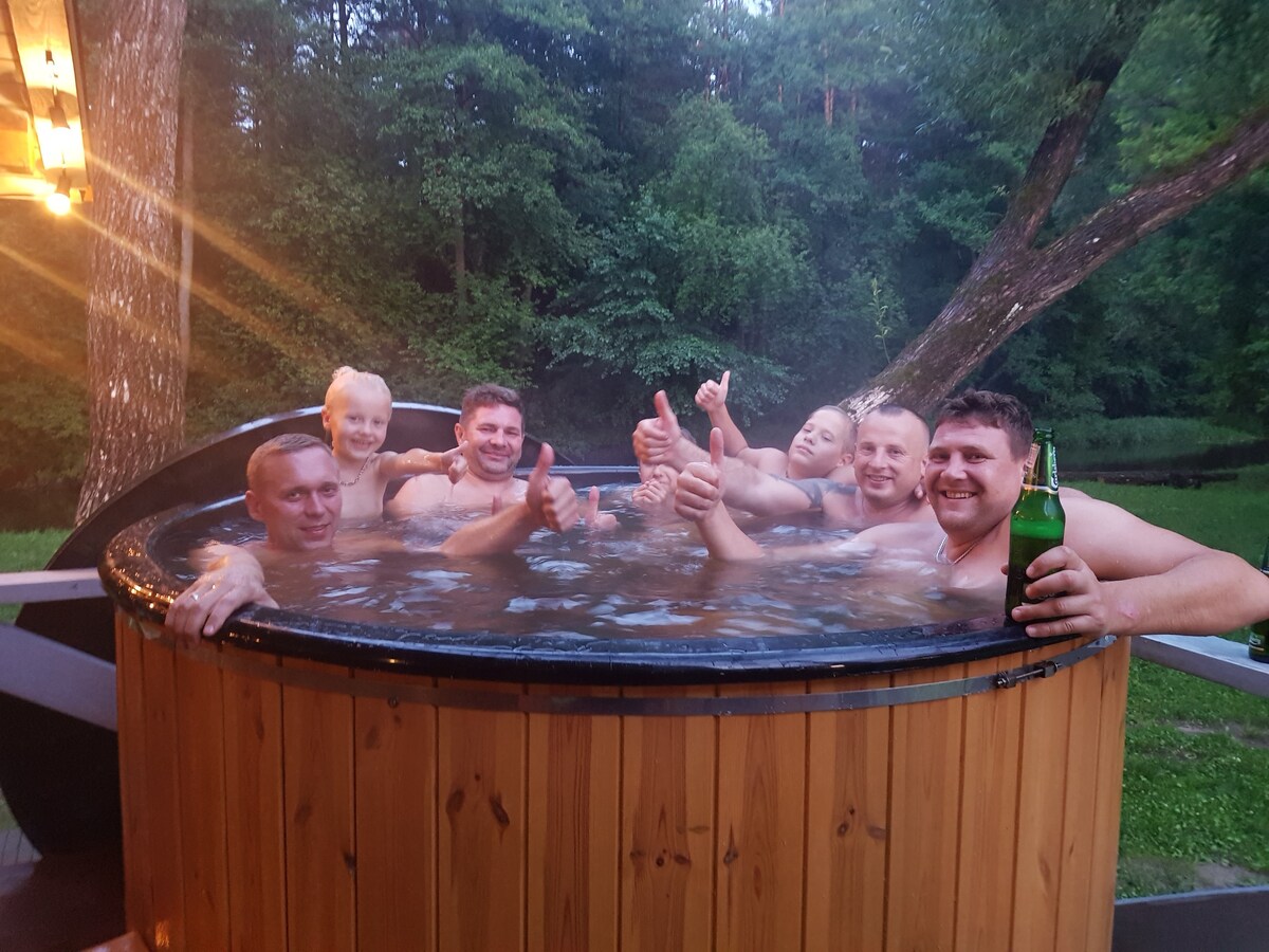 A wooden hot tub is surrounded by lush greenery, with steam rising gently from the water. Several individuals relax inside, enjoying the serene atmosphere, while one person holds a beverage. Soft lighting from nearby fixtures casts a warm glow over the scene.