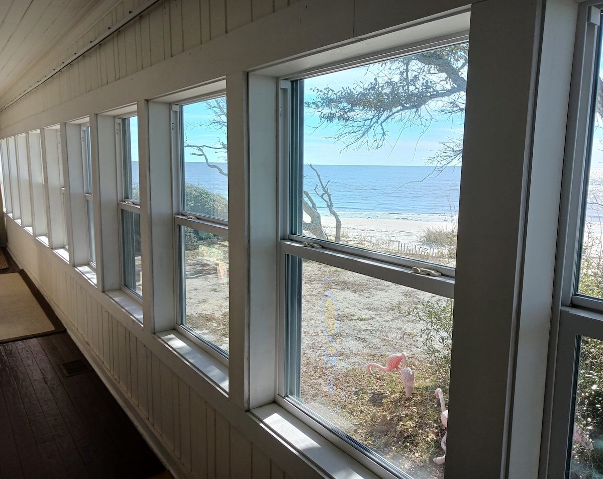 An ocean-facing sunroom is showcased with eleven windows, offering unobstructed views of the beach and water. Sunlight brightens the interior, highlighting the wooden floors and white paneling. Each window frames serene views of coastal vegetation and the tranquil shoreline beyond.