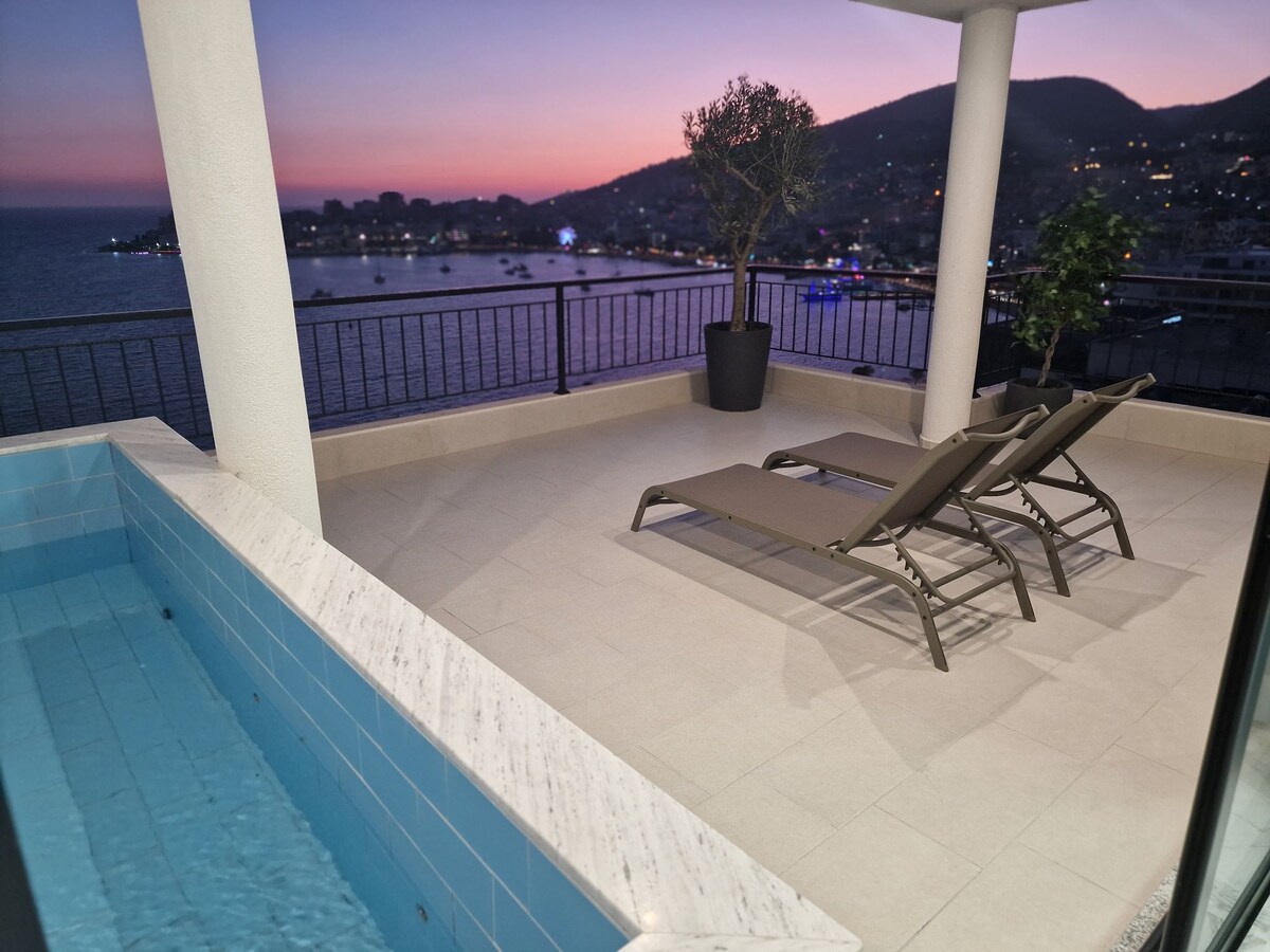 A private balcony offers stunning views of the sea and surrounding hills during sunset. Two lounge chairs are positioned beside a small pool, creating a serene space for relaxation. The soft glow of evening lights reflects off the water, enhancing the tranquil ambiance.