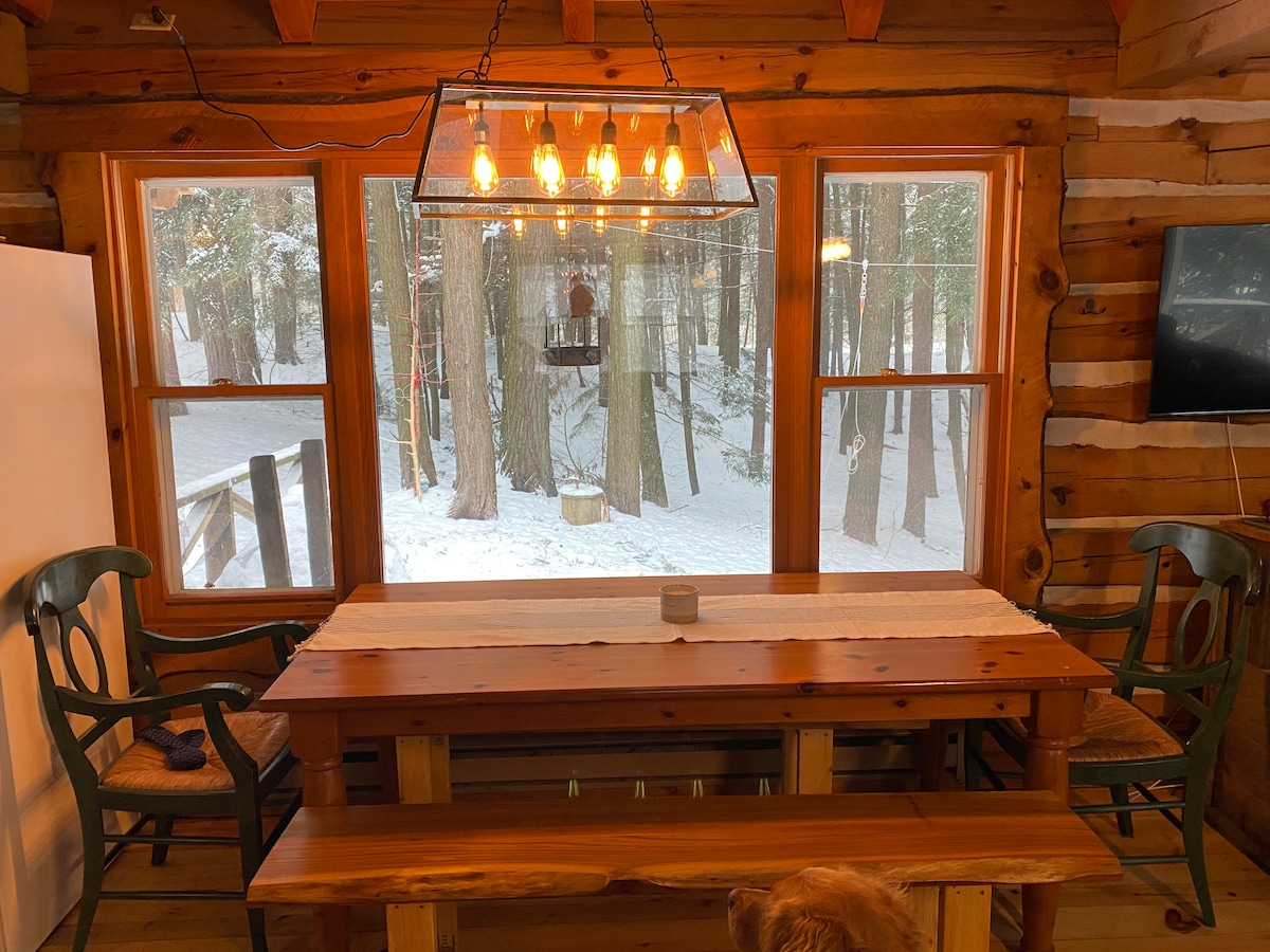 A dining area features a sturdy wooden table with a long bench and two green chairs. Large windows allow natural light and views of the snowy landscape outside. A light fixture with exposed bulbs hangs above, complementing the cabin's rustic decor.