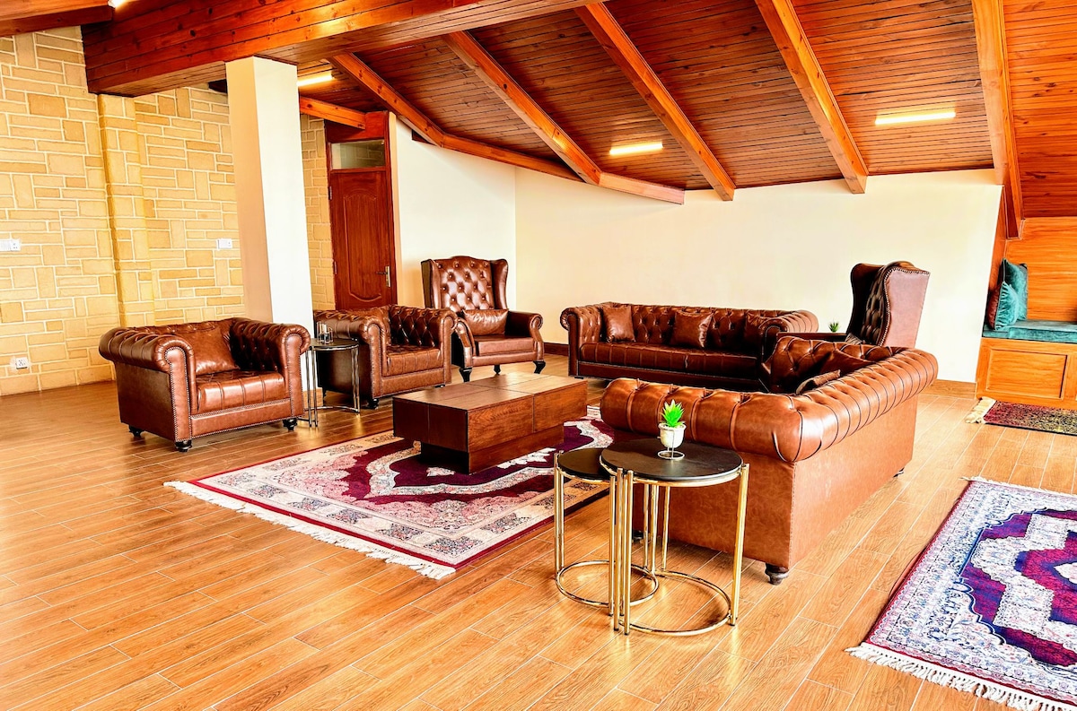 A spacious living area is highlighted by rich wooden beams and warm-toned furnishings. Two tufted leather sofas and a central coffee table are arranged on decorative rugs, while natural light fills the room through large windows, enhancing the inviting atmosphere.