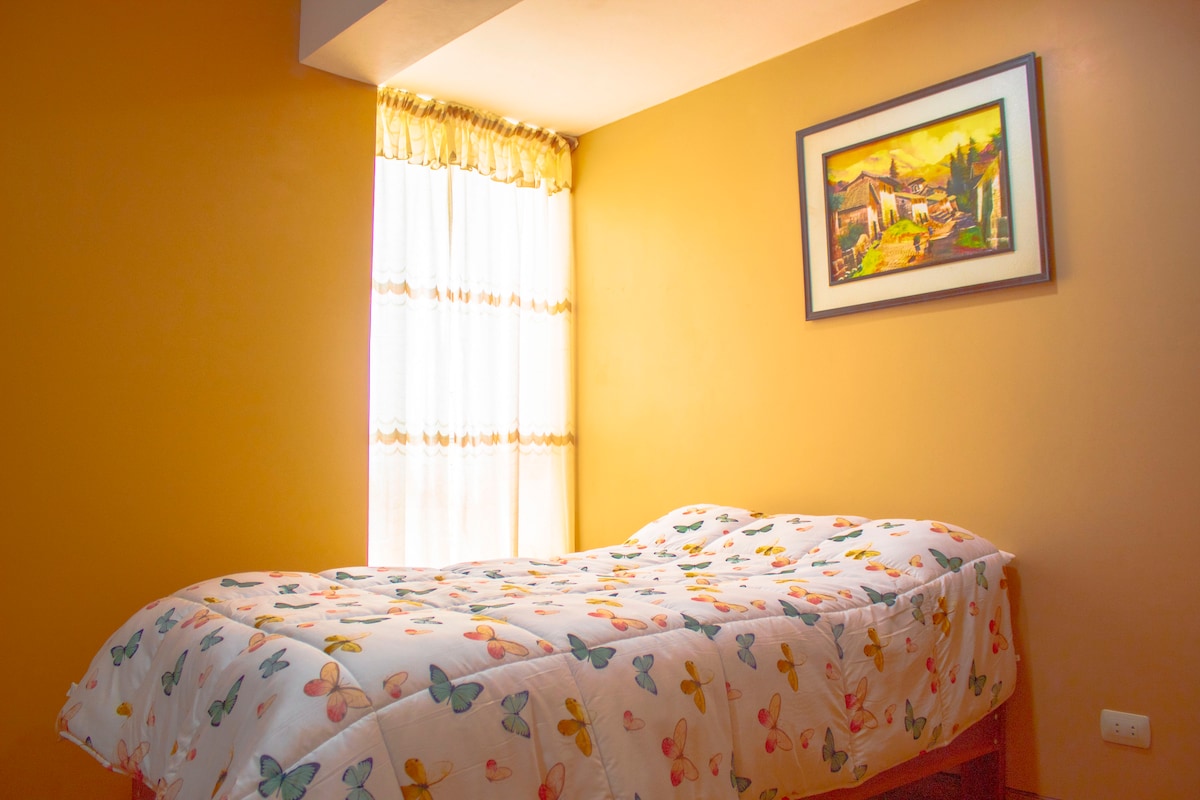 A well-lit sleeping area features a spacious bed adorned with a colorful butterfly-patterned blanket. Natural light filters through a window with sheer curtains, illuminating a warm, golden wall. A framed artwork adds a touch of character to the space.