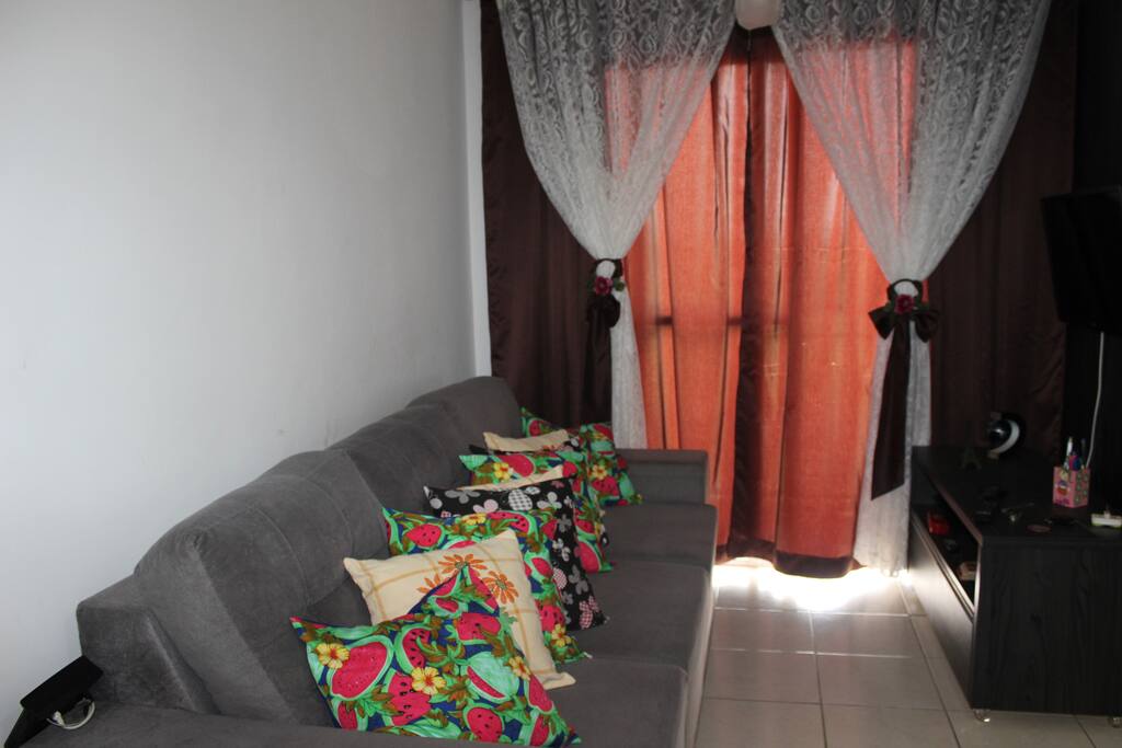 Sala de estar com televisão, sofá-cama retrátil de casal, cortinas, espaço para um colchão de solteiro e mesa de jantar.