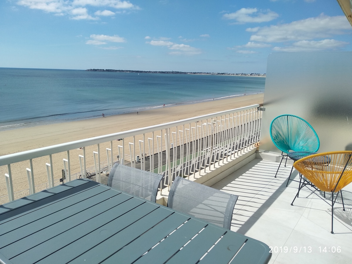 A spacious terrace offers outdoor seating with a dining table and chairs while framed by a stunning view of the beach and ocean. The calm waters are complemented by sandy shores and a clear blue sky, creating an inviting space for relaxation.