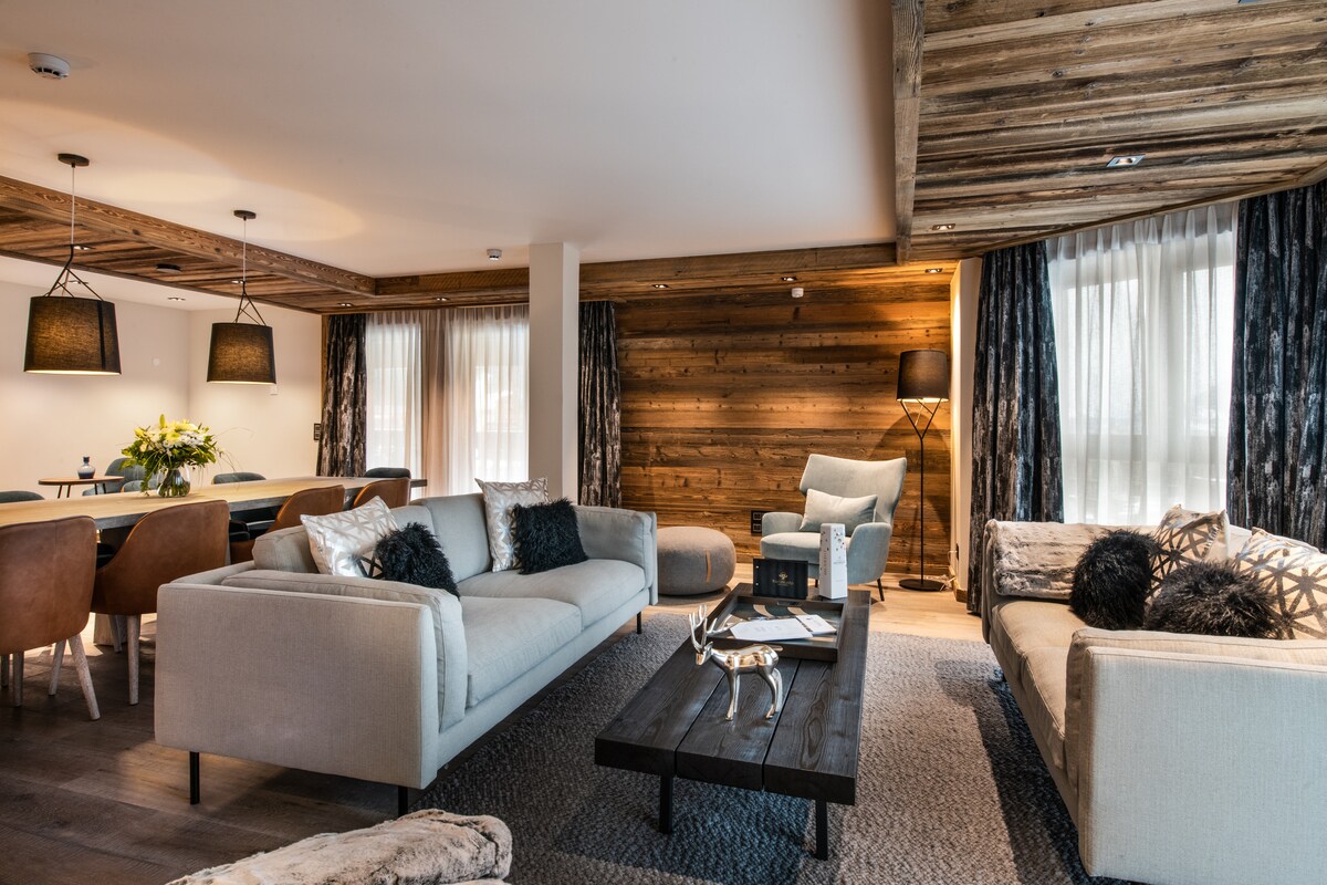 A spacious living area features a comfortable seating arrangement with two light-colored sofas and decorative pillows. A dark wooden coffee table sits centrally, complemented by a textured rug. Large windows with sheer curtains allow natural light to illuminate the room, while wooden accents add warmth.