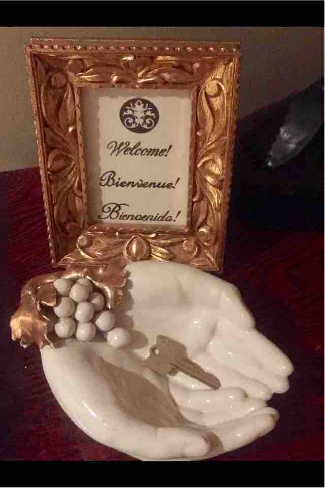A decorative frame displays multilingual welcome messages against a textured background. Below, a ceramic hand is positioned to hold a key and a cluster of grape-like ornaments, emphasizing hospitality and charm in a thoughtful arrangement.