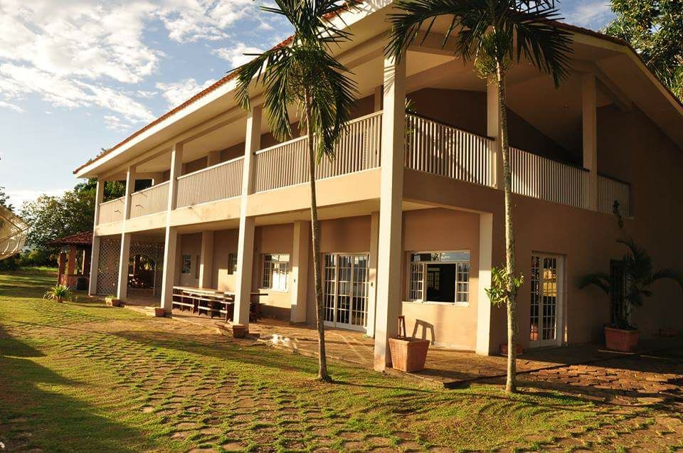 The exterior of the ranch is shown, highlighting a two-story building with a broad balcony supported by columns. Windows with white frames reflect natural light, while the surrounding grassy area features a stone pathway. Palm trees add greenery to the tranquil setting.