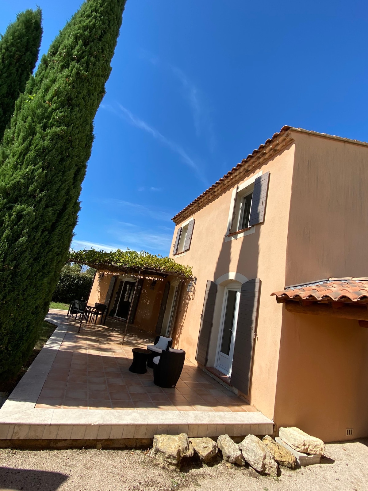 The exterior of a villa is shown, featuring a warm-toned stucco façade and terracotta roof. A covered terrace with seating overlooks a well-maintained garden. Tall cypress trees frame the property under a clear blue sky, creating a serene ambiance in the surrounding area.