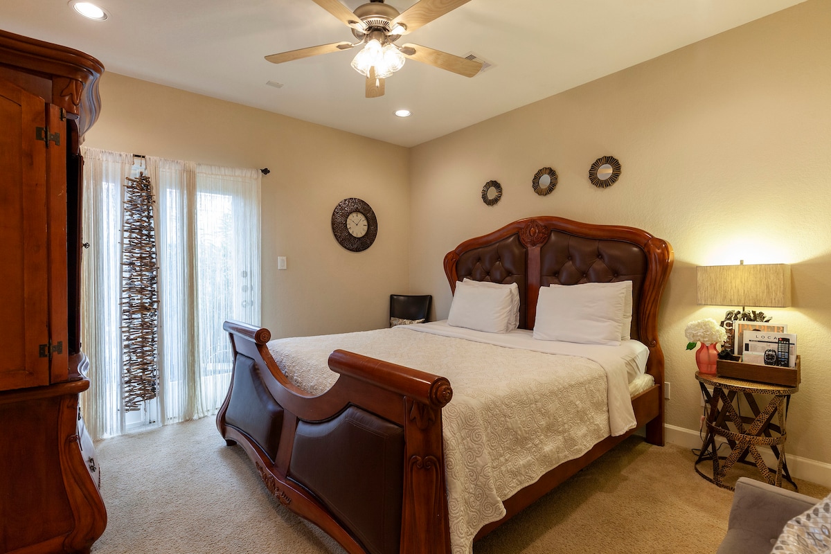 A comfortable bedroom features a dark wood bed frame with a padded headboard and light bedding. A ceiling fan and bedside lamp provide ambient lighting. Windows adorned with sheer curtains create a bright atmosphere, complemented by decorative wall accents. A wooden wardrobe is visible in the corner.