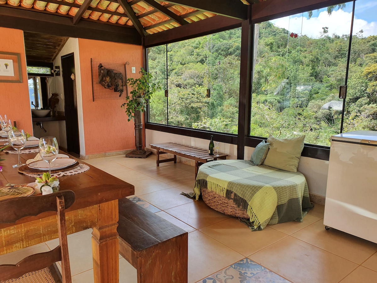 A bright living area features expansive glass panels that open to the lush surrounding landscape. A rustic wooden dining table is set for a meal, while a cozy seating area with a woven blanket invites relaxation. A small refrigerator is positioned nearby.