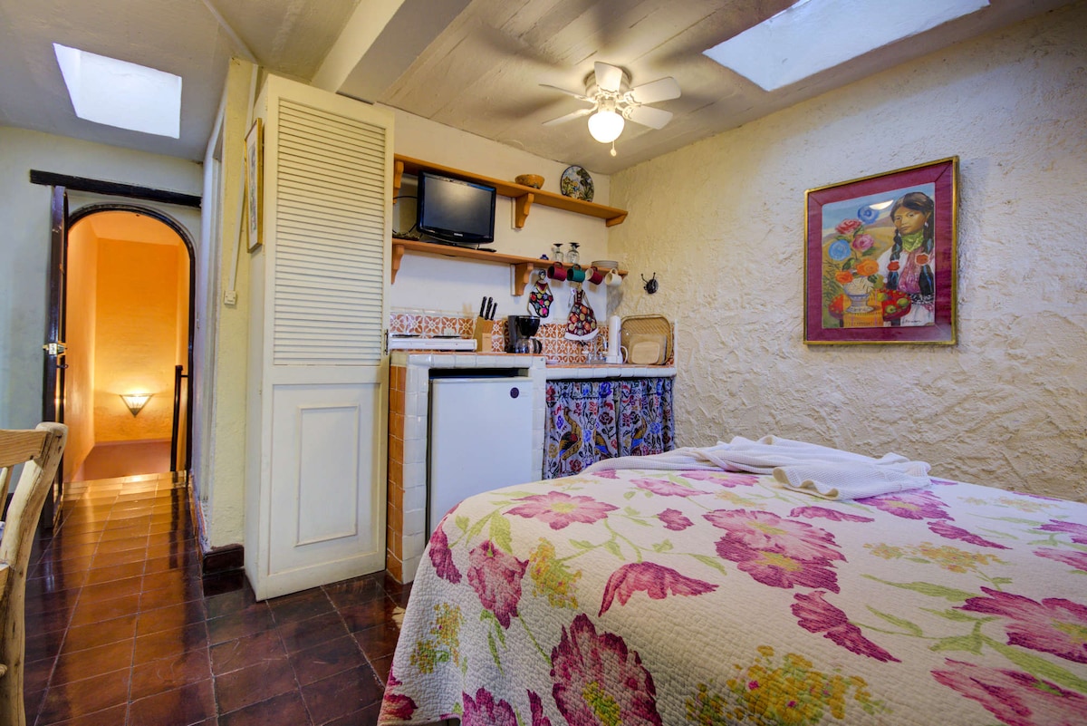 The studio features a cozy queen-sized bed adorned with a floral quilt. A small kitchenette equipped with a refrigerator and cooking essentials sits nearby, with a ceiling fan overhead providing airflow. Natural light enters through the skylight, illuminating the light-colored walls and rustic decor.