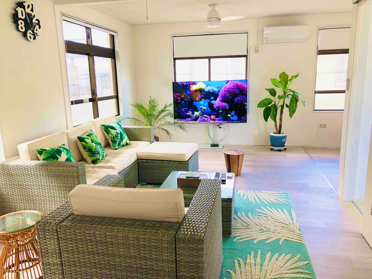 A modern living room features a comfortable seating area with a sectional sofa adorned with green cushions. A large aquarium provides a colorful backdrop, while houseplants enhance the natural ambiance. Natural light fills the space through multiple windows.