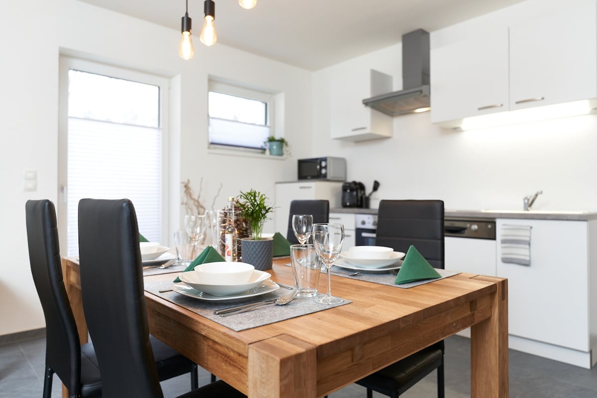 A dining area features a wooden table set for six with neutral dinnerware and green napkins. Black chairs surround the table, and a small plant adds a touch of greenery. A modern kitchen with white cabinetry and stainless appliances is visible in the background.