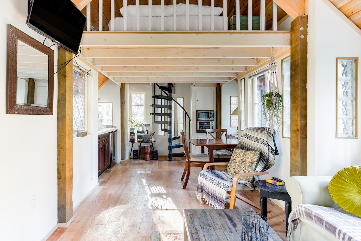 The living area features a mix of wood and white walls with ample natural light streaming through multiple windows. A spiral staircase leads to the loft bedroom. A dining table and a comfortable armchair are visible, along with a large TV mounted on the wall.