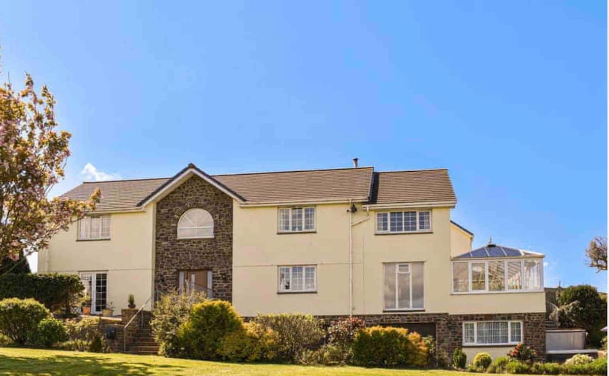 The spacious exterior of the house is showcased, highlighting its dual textures of stone and stucco. A conservatory with large windows adds a bright feature to one side, complementing the surrounding landscaped garden with lush greenery and colorful flowers under a clear blue sky.