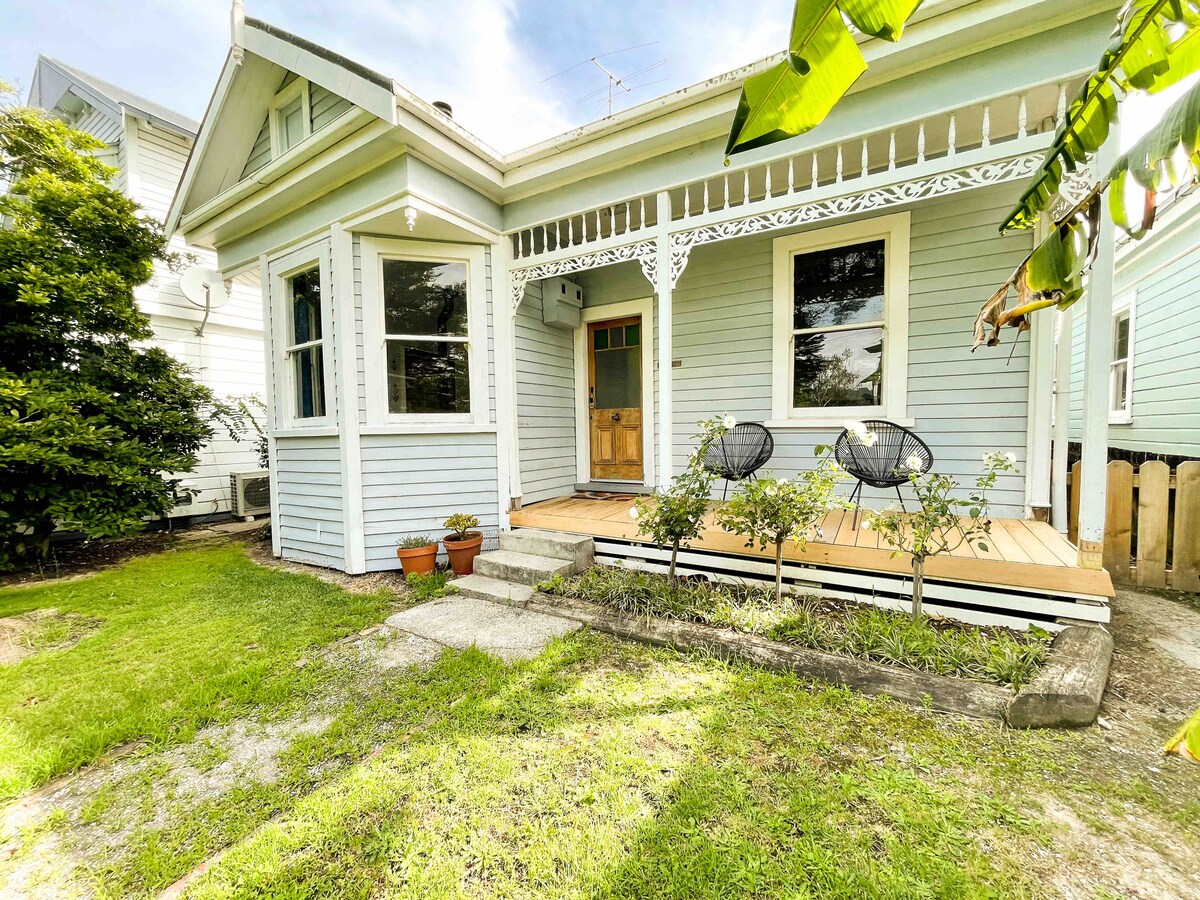 The exterior of a charming villa is shown, featuring a wooden porch with two black chairs on a wooden deck. Lush green grass surrounds the house, complemented by potted plants and decorative details on the eaves. Large windows allow natural light to enhance the inviting facade.
