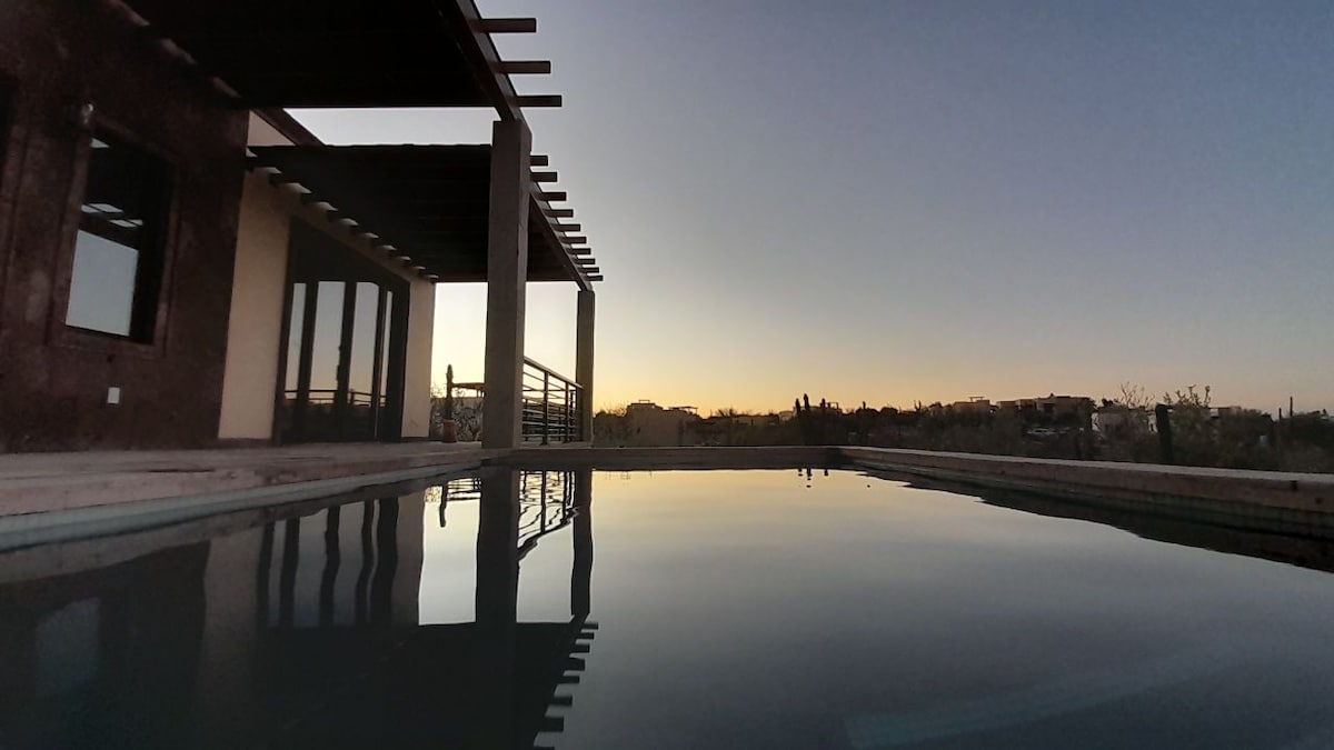 A tranquil pool reflects the silhouette of a rooftop terrace at dusk, with gentle ripples on its surface. The surrounding area is framed by a modern structure, partially obscured by the soft glow of a setting sun on the horizon.
