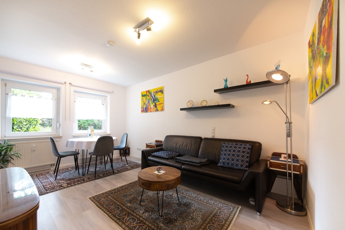 A welcoming living area features a dark sofa with decorative cushions, a light wooden coffee table, and a medium-sized dining table surrounded by four chairs. The room is illuminated by ceiling lights and natural light from two windows, with colorful artwork on the walls.