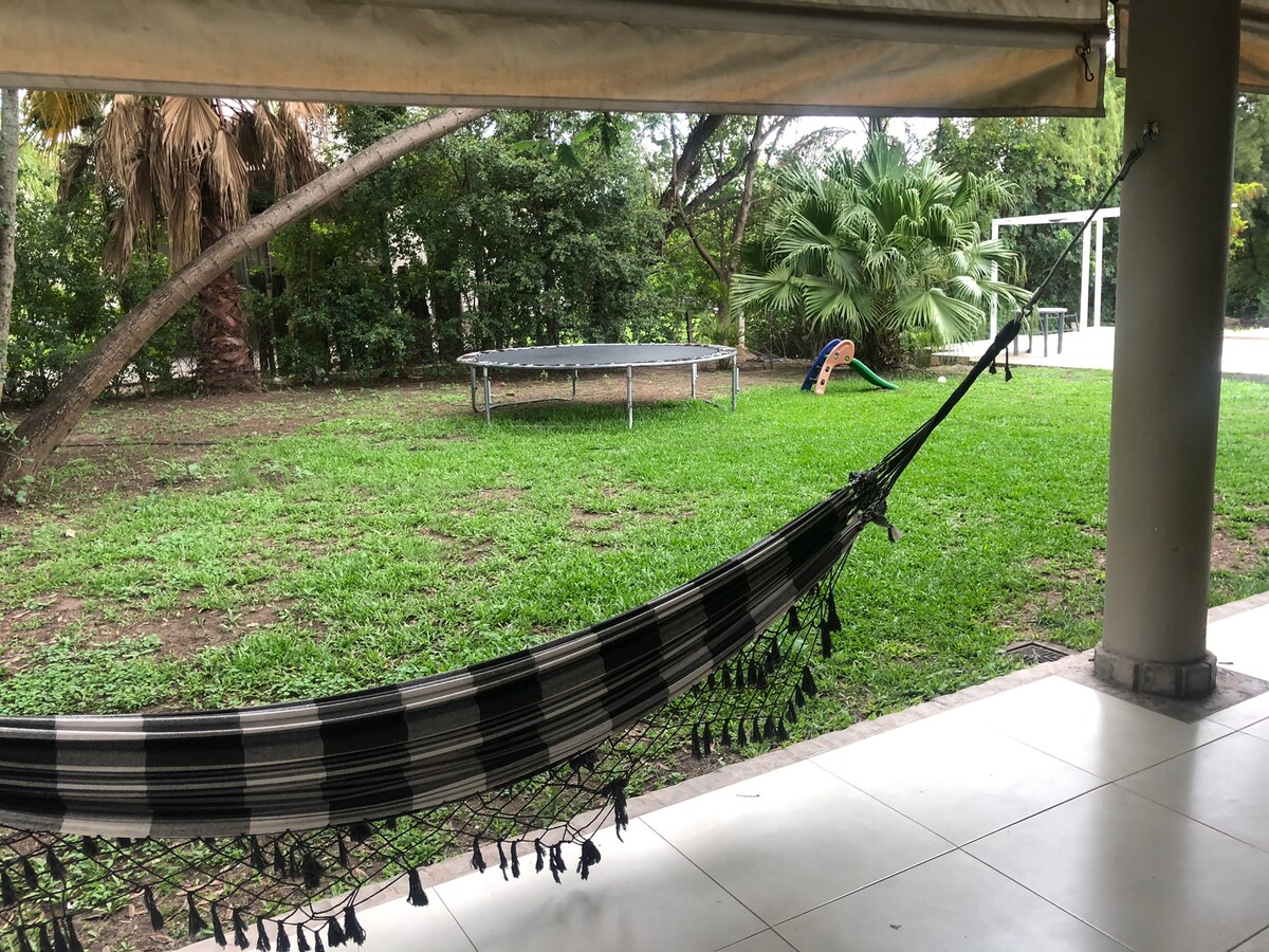 A spacious outdoor area is showcased, featuring a green lawn surrounded by trees. A hammock is suspended under a shelter, creating a relaxing spot. In the background, a trampoline and a play structure are visible, inviting for leisure activities.