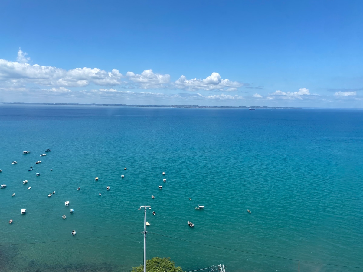 A panoramic view of the ocean is presented, featuring a vibrant blue sea dotted with several small boats. The horizon is visible in the distance, framed by a clear sky with a few scattered clouds, creating a serene atmosphere.
